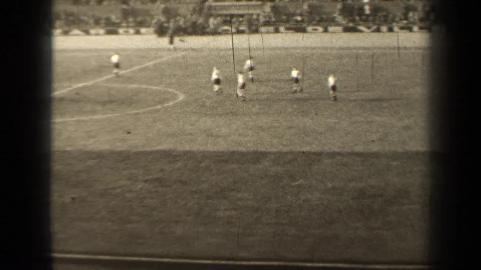 1947 Paris 8mm frame showing distant soccer players spread across a marked grass pitch, with spectators faintly visible beyond the field.