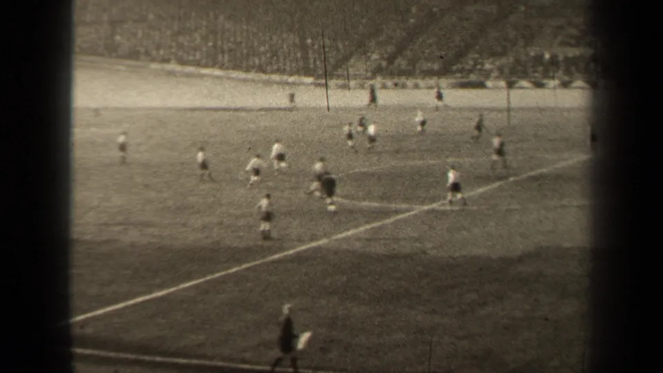 A distant 8mm view shows football players moving across the Paris pitch, with stands and a sideline area behind them.