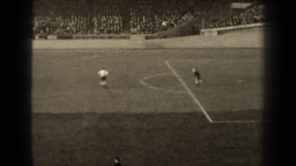 Late-frame view of the 1947 Paris football match near the penalty area, with distant players and spectators in the stands.
