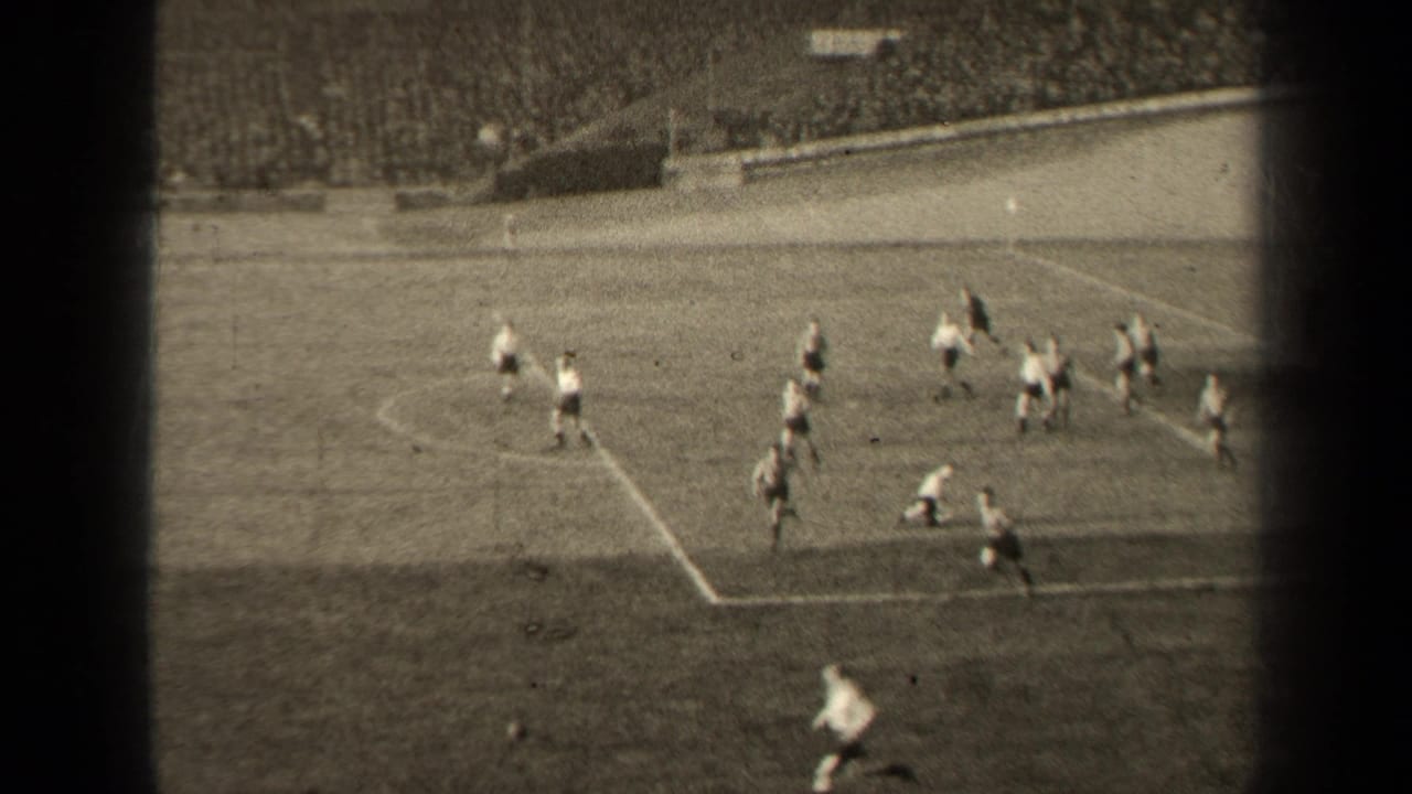 Soccer players on a grass field near the penalty area with crowded stands behind them.