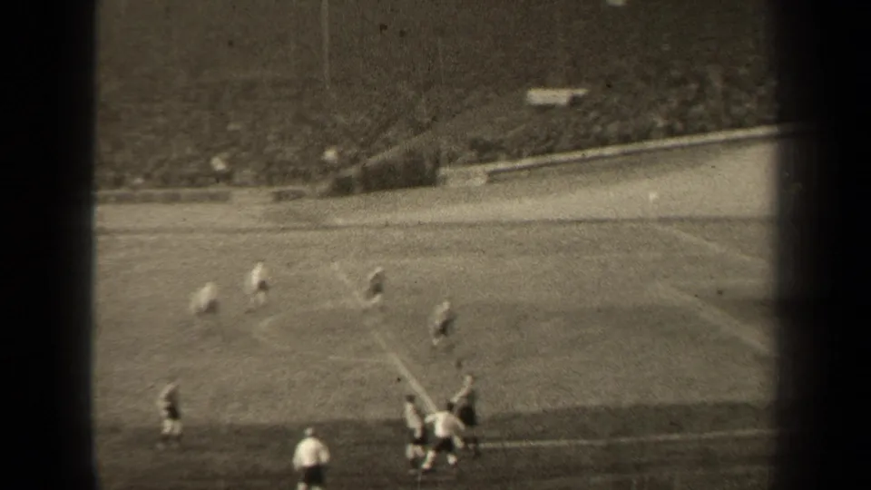 Distant 8mm frame from a Paris soccer match, with players clustered near the sideline and stadium seating in the background.