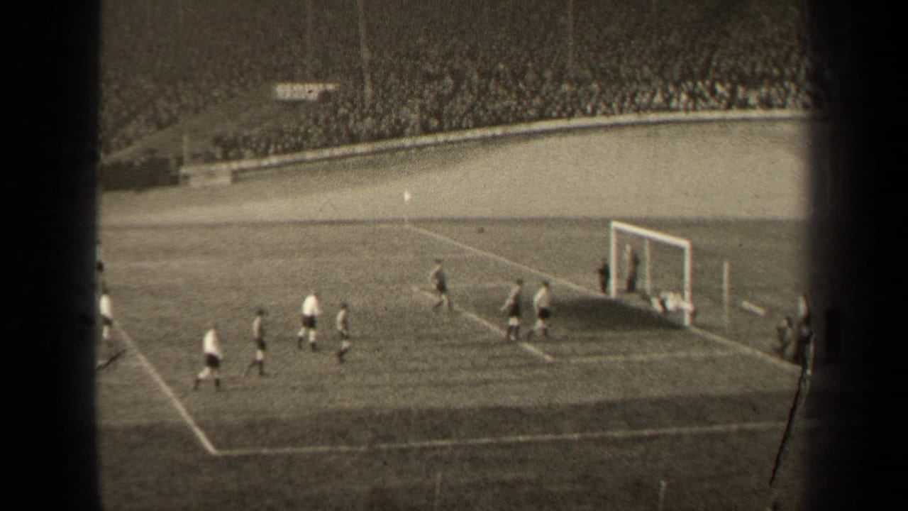 Football players gather near a goal on a grass pitch with spectators in the stands.