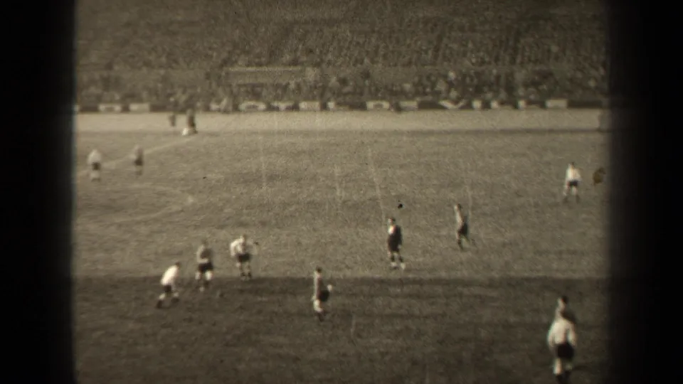 Wide 1947 Paris soccer match frame showing players, a dark-clothed official, open pitch, and crowded stadium seating.