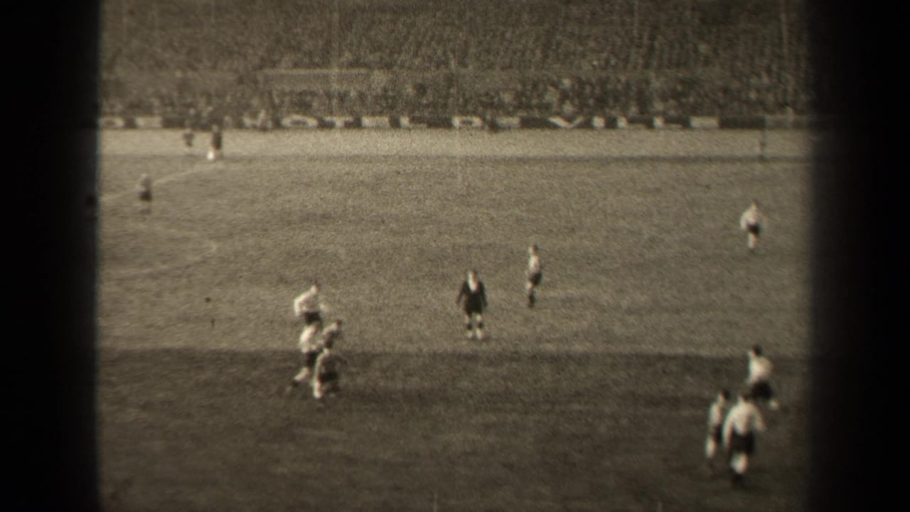 Football players spread across a grass pitch with crowded stands beyond.