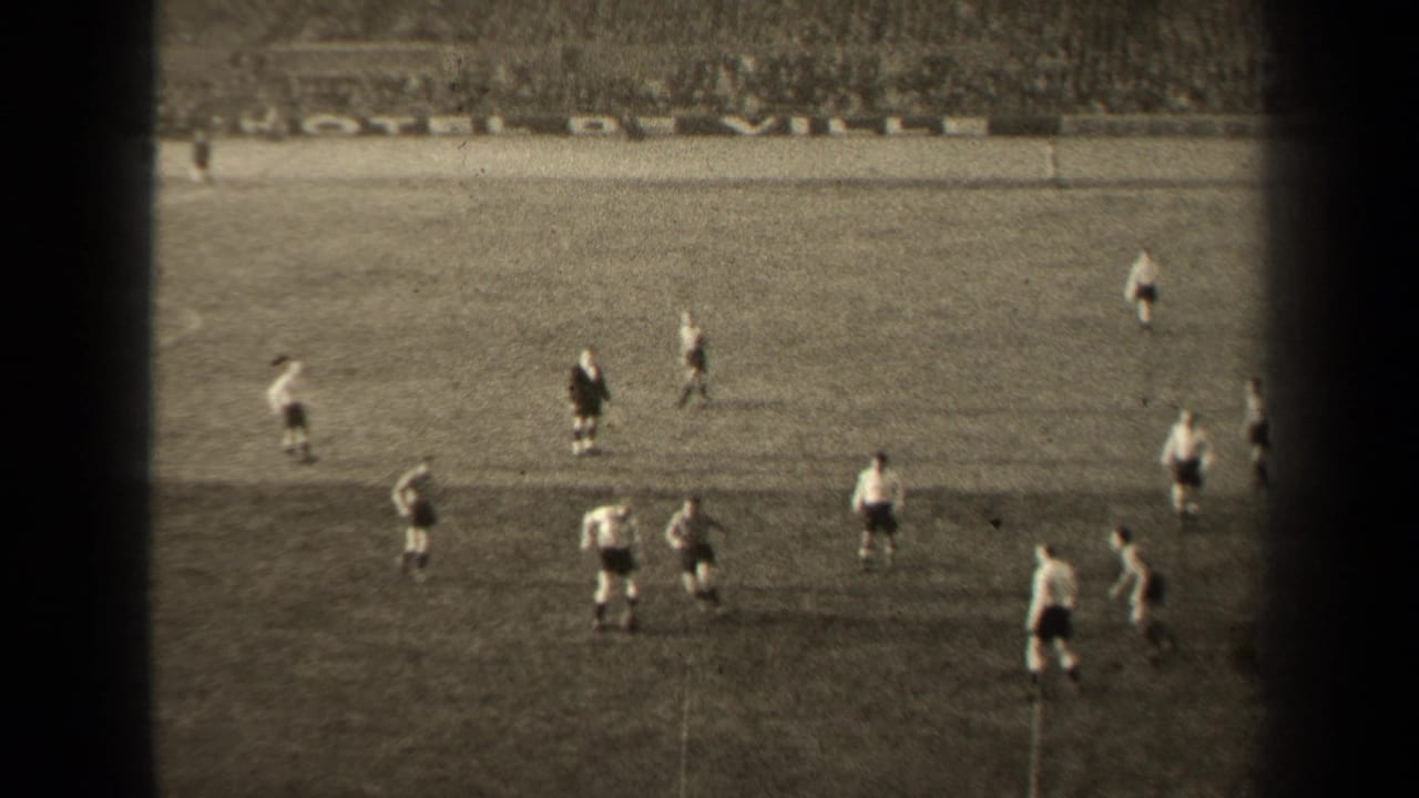 High-angle 8mm frame of a Paris football match, showing players positioned around the ball on the grass pitch.