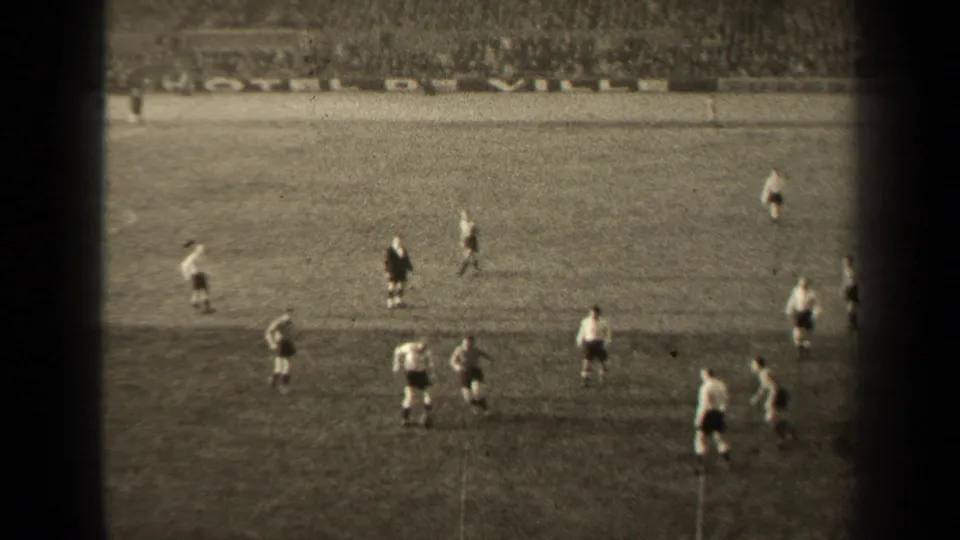 High-angle 8mm frame of a Paris football match, showing players positioned around the ball on the grass pitch.