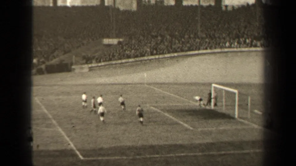 Football action near the goalmouth in the supplied 1947 Paris match, with several players approaching the net under stadium stands.