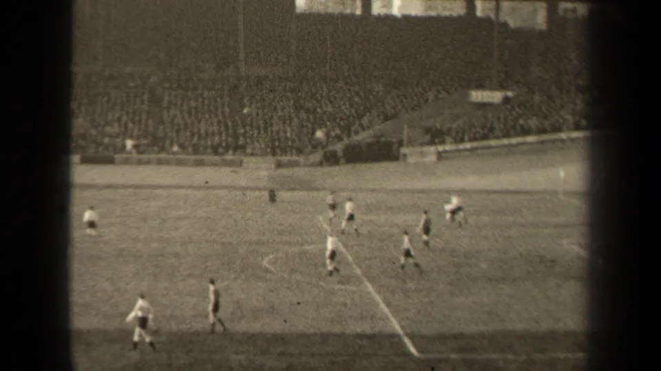 Black-and-white 8mm frame from a 1947 Paris football match, with players spaced across the pitch below packed stands.
