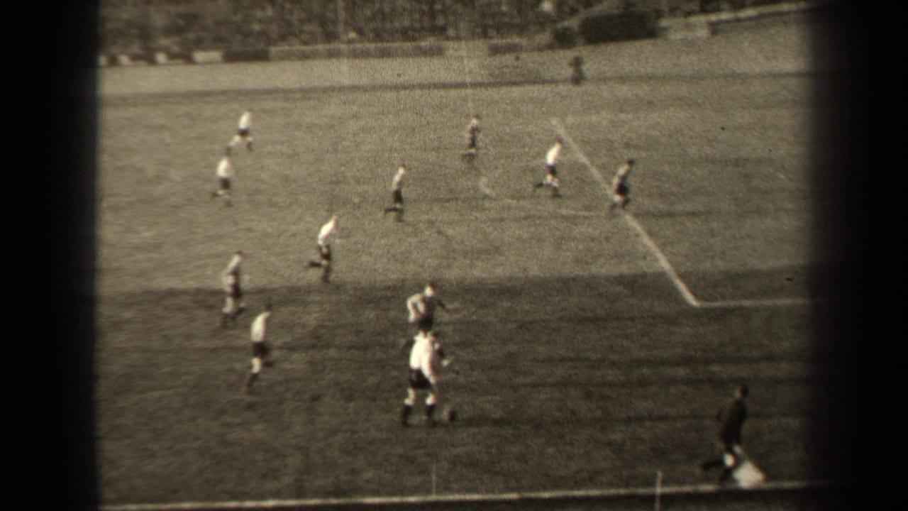 Soccer players cluster near the touchline with a packed stadium crowd beyond the field.
