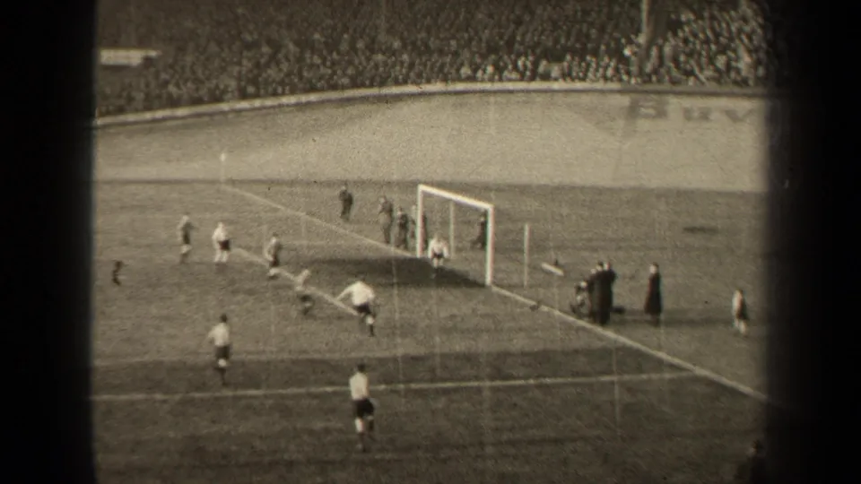 Distant 1947 Paris match frame showing players near the goalmouth, sideline figures, and a large stadium crowd beyond the pitch.