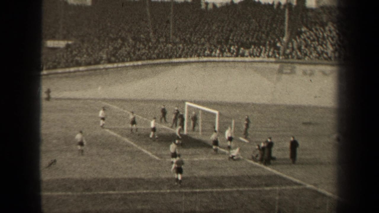 A goalmouth play unfolds with several football players and a packed stand behind them.