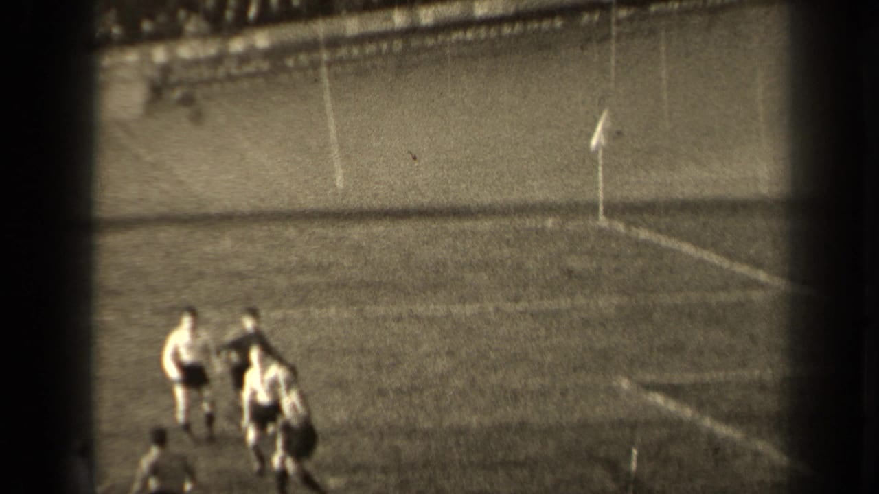 Adult male players contest the ball near the corner area of a Paris soccer pitch, with a flag and spectators beyond the field.