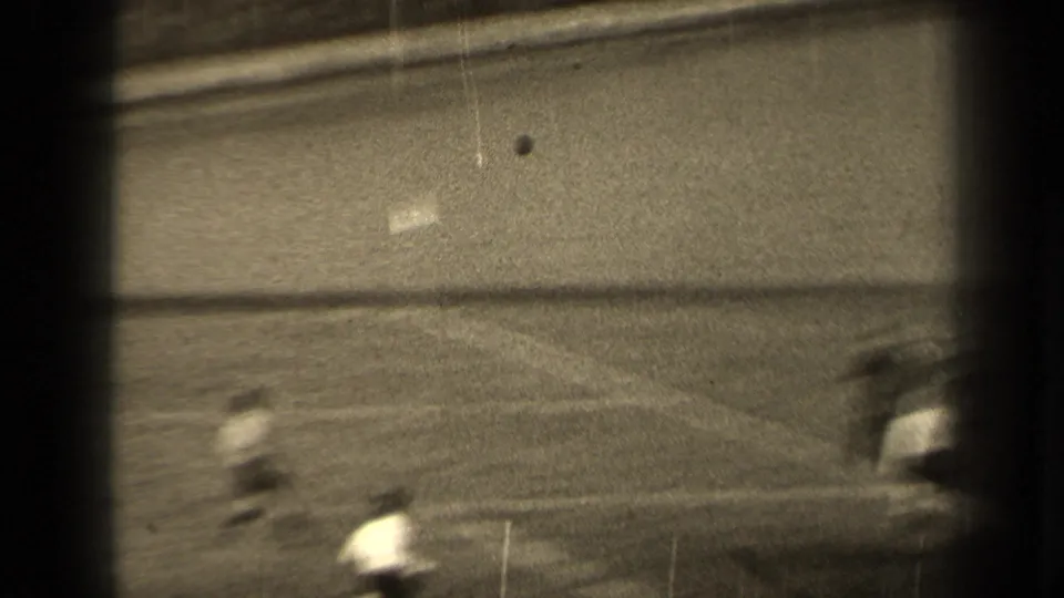 A spectator-shot 8mm view in Paris shows blurred football players running across the field behind dark foreground obstructions.