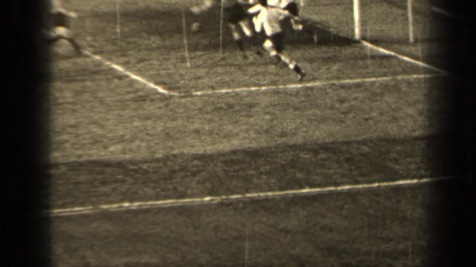 Players gather near the goal area on a Paris soccer pitch, with white markings, a partial goal structure and grainy 8mm texture.