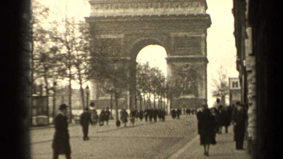 1947 Paris home-movie frame showing pedestrians on a city sidewalk with a large stone arch visible in the distance.