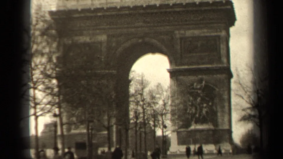 The Paris monument fills the frame, with the arch opening, facade relief, bare branches, and distant visitors visible below.