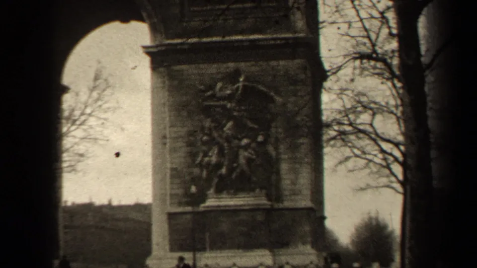 Closer 8mm frame of the Arc de Triomphe side relief in Paris, with bare branches, dark film edge, and a few pedestrians below.