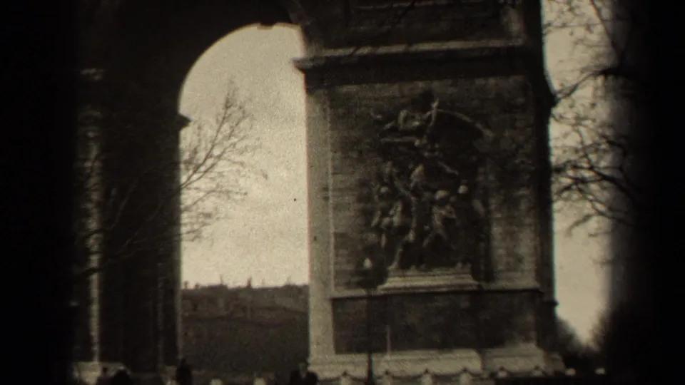 A low-angle Paris home-movie frame shows the Arc de Triomphe relief panel, arch edge, bare trees, and distant figures near the base.