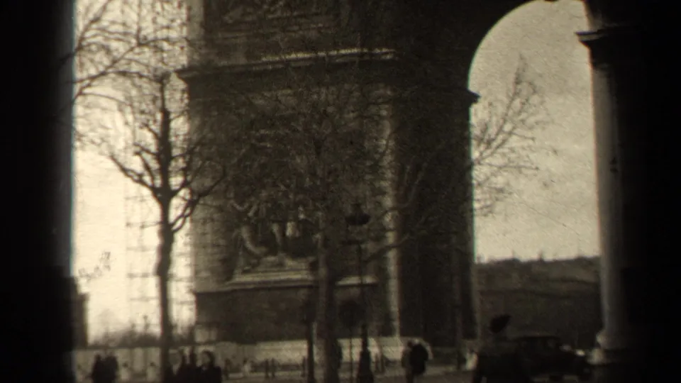 1947 Paris home-movie frame showing the Arc de Triomphe partly obscured by a bare tree, with pedestrians and a dark car near the base.