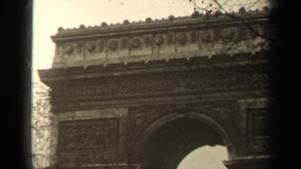 1947 Paris frame showing the Arc de Triomphe upper facade, arch opening, bare trees, cloudy sky, and soft 8mm texture.