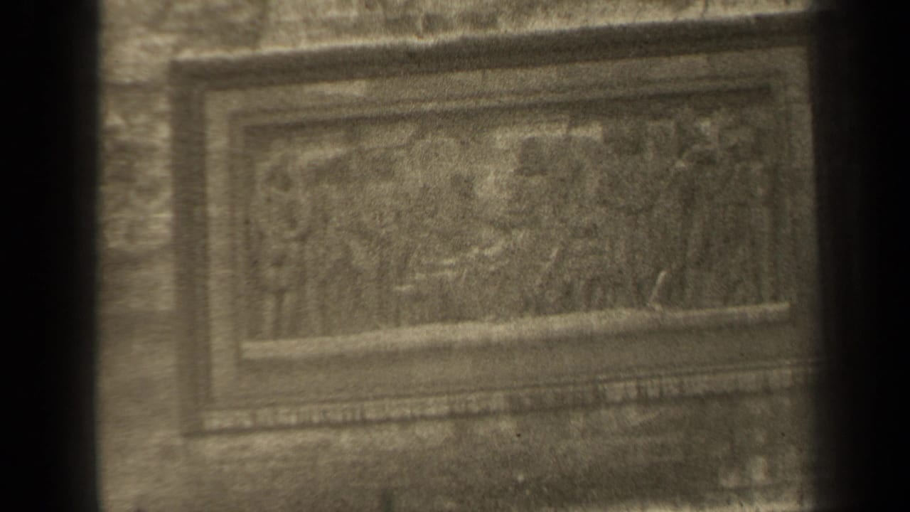 1947 Paris home-movie frame showing a tarnished rectangular bas-relief with sculpted figures, seen through dark vertical obstructions.