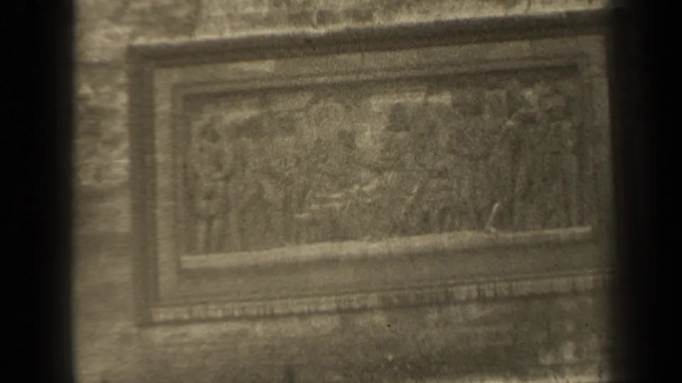 1947 Paris home-movie frame showing a tarnished rectangular bas-relief with sculpted figures, seen through dark vertical obstructions.