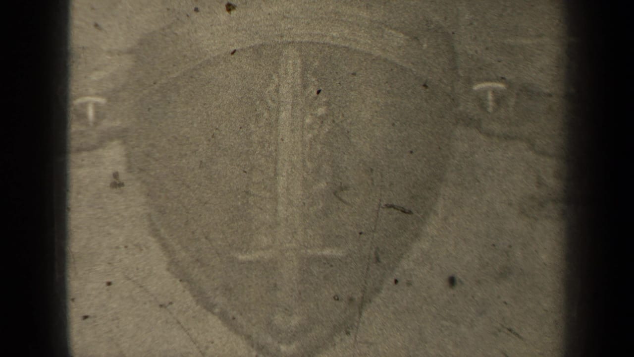 Close view of a shield-like emblem with a sword shape and letter-like mark on worn film.