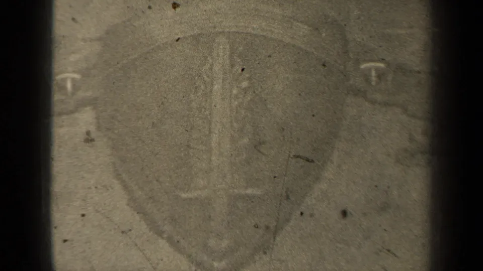 1947 8mm frame showing a shield-like emblem with a sword shape and letter-like mark, softened by grain, dust and dark gate edges.