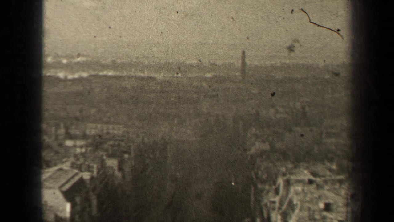 Hazy high-angle city view with rooftops, trees and a distant spire.