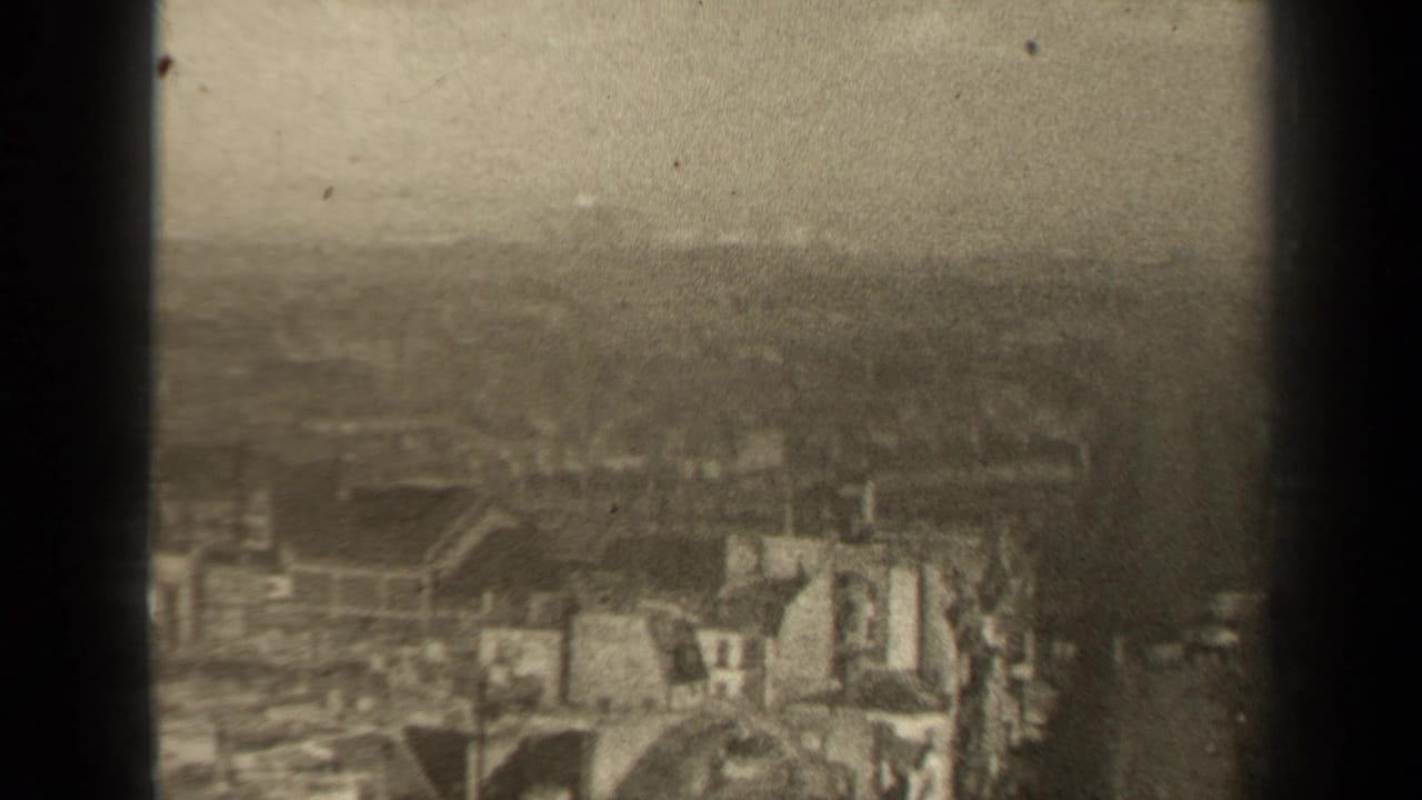 Soft cityscape view with rooftops, pale facades, haze, and dark vertical foreground bars.