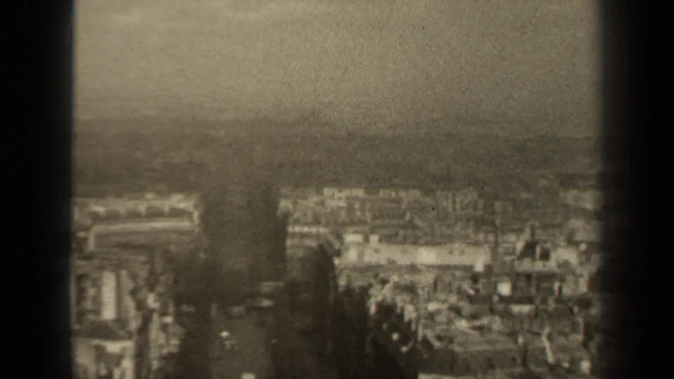 1947 Paris home-movie frame with rooftops, a central street corridor, haze, and heavy 8mm grain.