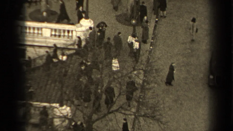 1947 Paris street frame showing people walking naturally across a cobblestone area, with bare branches, a light stone balustrade, and soft 8mm grain.