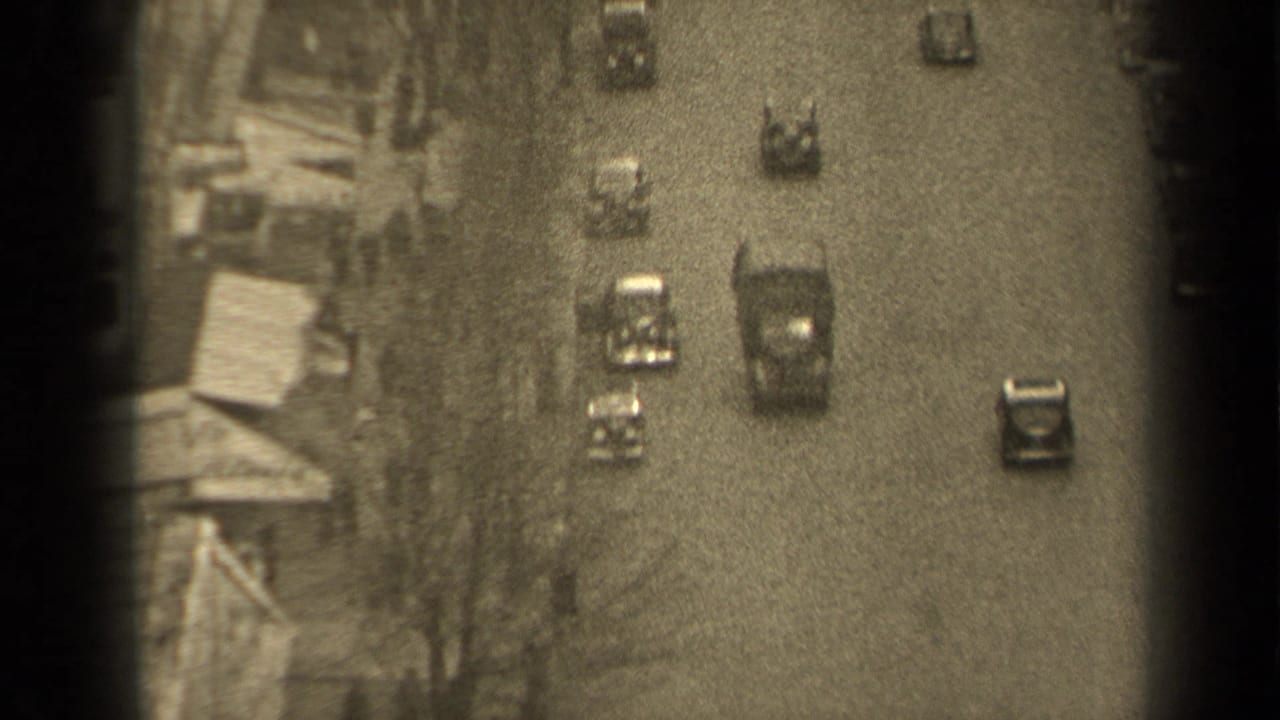 High-angle 1947 Paris street view with mid-century cars moving along a wide road beside building fronts and sidewalks.