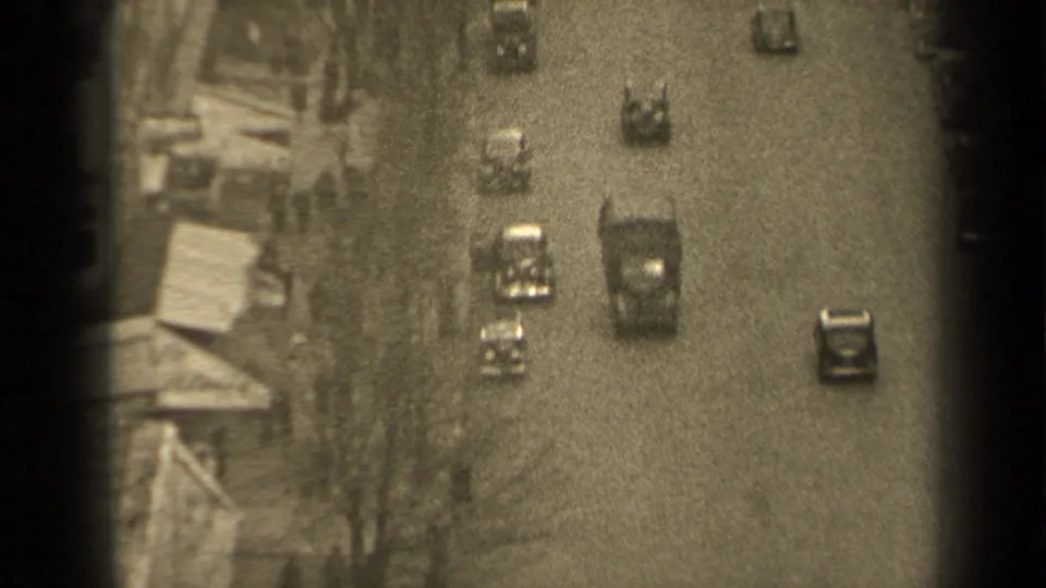 High-angle 1947 Paris street view with mid-century cars moving along a wide road beside building fronts and sidewalks.