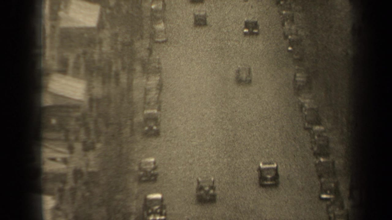 Cars travel on a wide street with parked vehicles and buildings along the side.