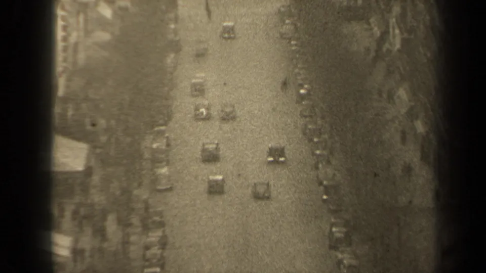 Grainy 8mm frame of a 1947 Paris street, with cars moving away below and buildings framing the roadway.