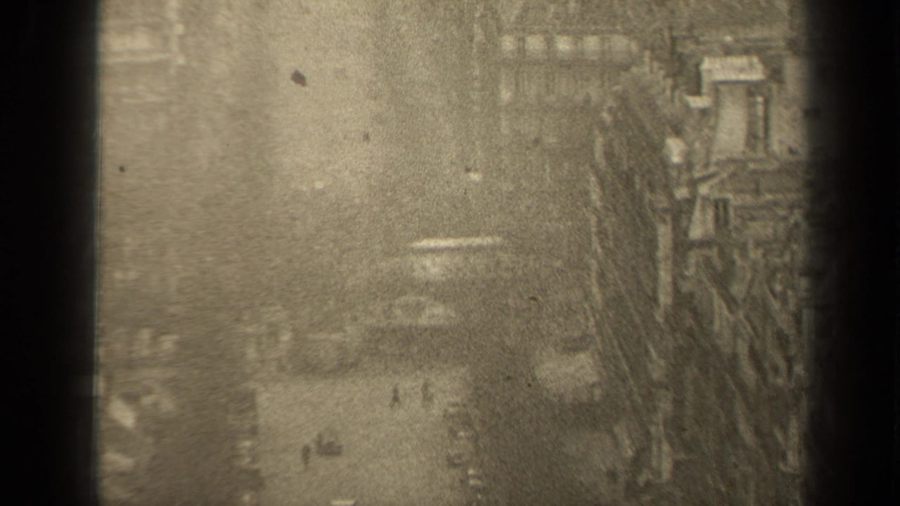 Upper-window view of a city street with cars, pedestrians, bare trees, and dense building fronts.