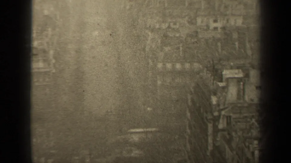 Grainy 8mm frame of a Paris street from above, showing buildings, bare trees, small vehicles, and a dark vertical edge in the foreground.