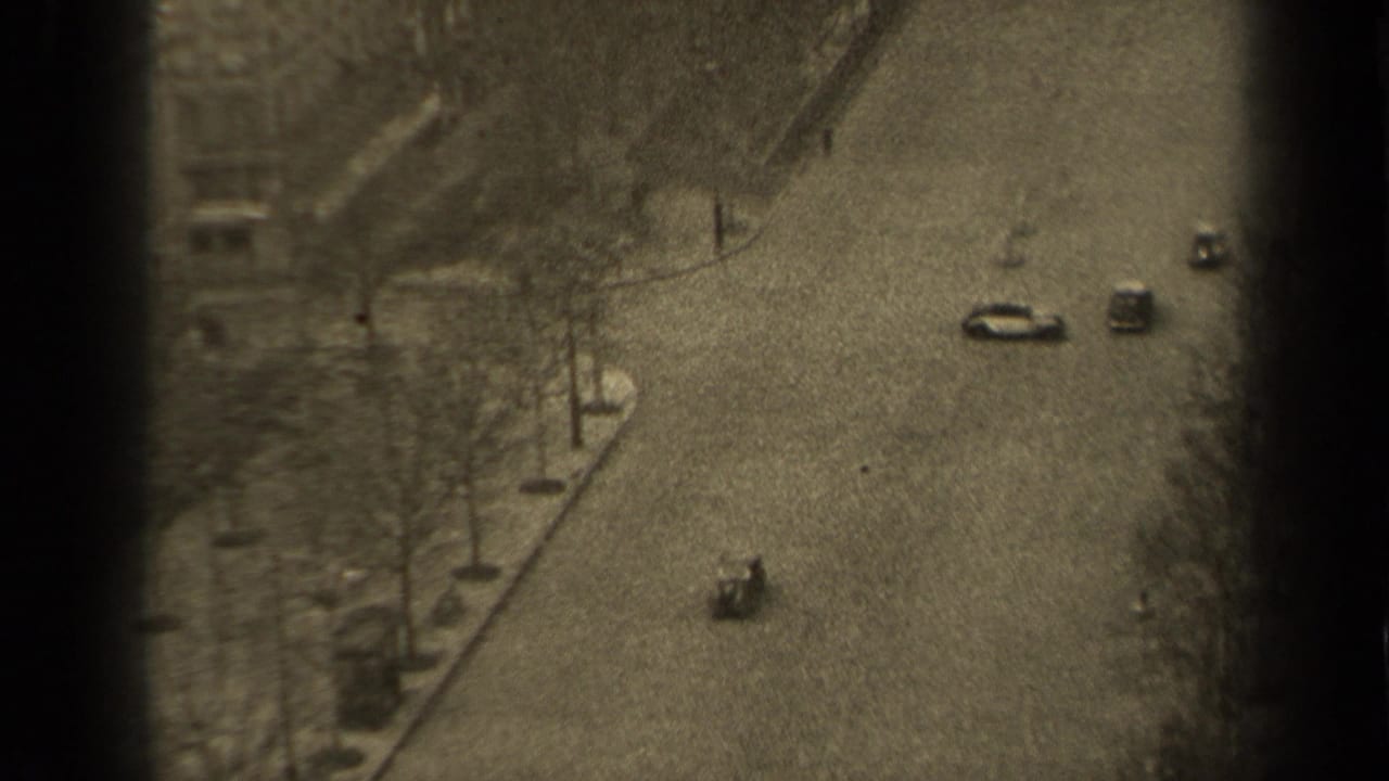1947 Paris home-movie frame showing a high-angle street view with cars, bare trees, sidewalks, and distant pedestrians.