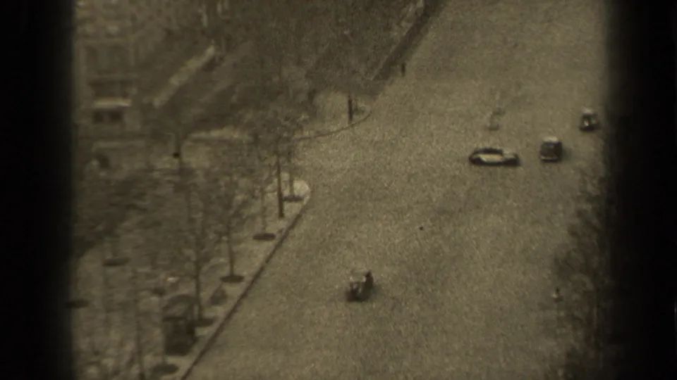 1947 Paris home-movie frame showing a high-angle street view with cars, bare trees, sidewalks, and distant pedestrians.