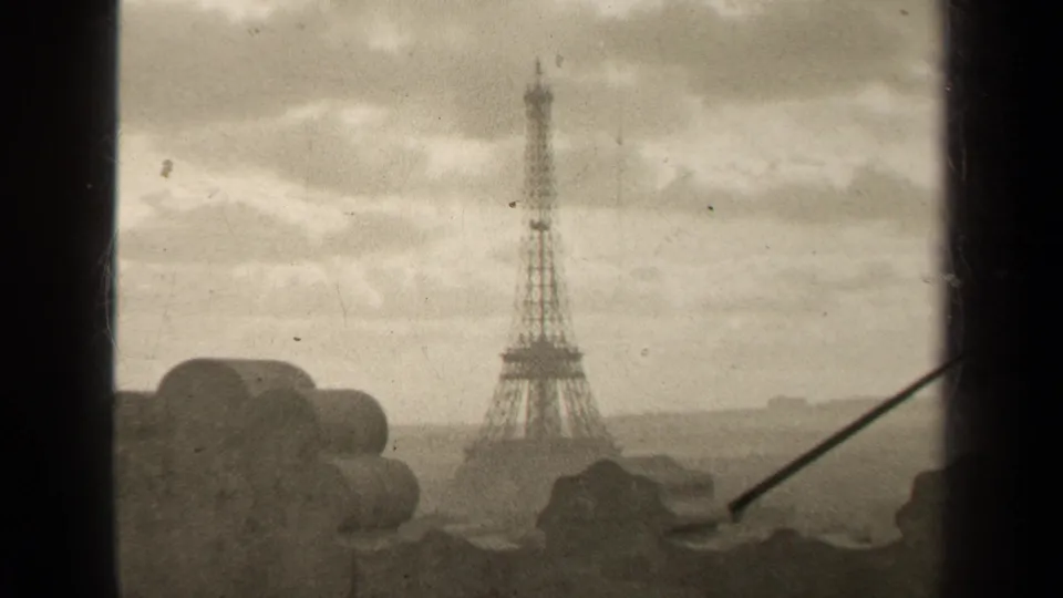 1947 Paris home-movie frame showing the Eiffel Tower through a high window, with cloudy sky and a distant city horizon.