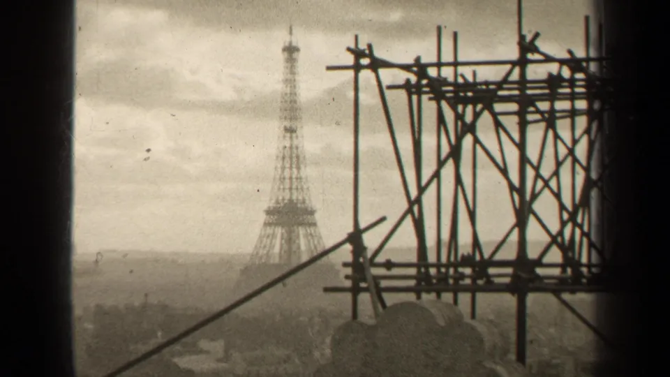 Distant Eiffel Tower in Paris is framed by dark scaffolding, with soft film grain and a muted skyline below.
