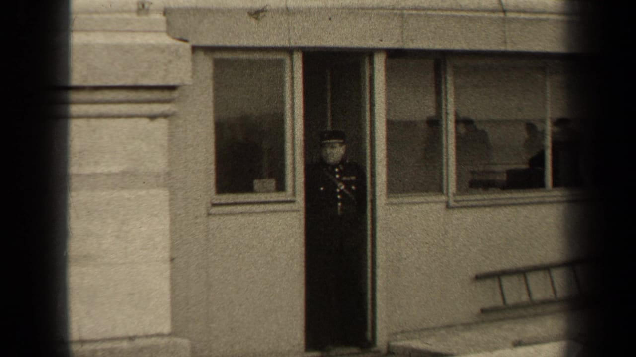 Uniformed man stands in a doorway beside windows on a stone building facade.