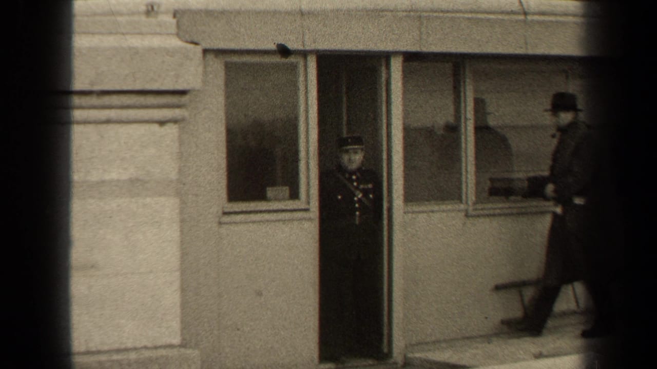 Uniformed man stands in a doorway as a civilian in a long coat approaches outside a windowed building.