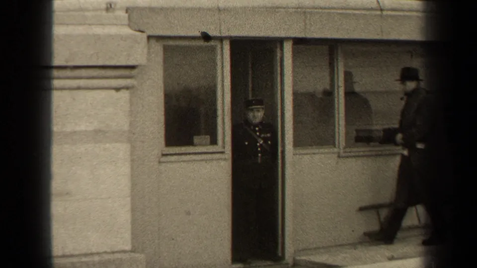 In 1947 Paris, a uniformed man stands at a building doorway while a civilian in a long coat and hat walks nearby.