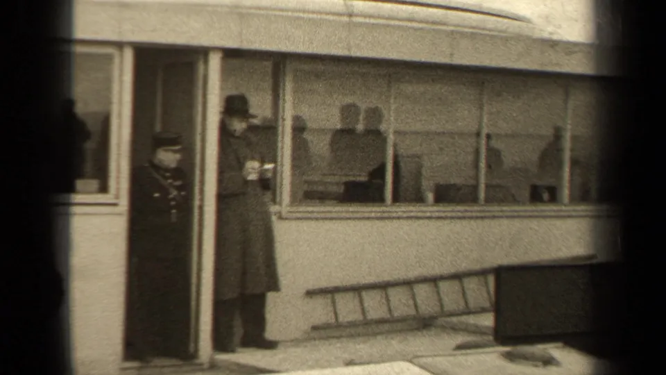 A civilian in a long coat and hat stands near the windows of a Paris building, with a uniformed doorway figure nearby.