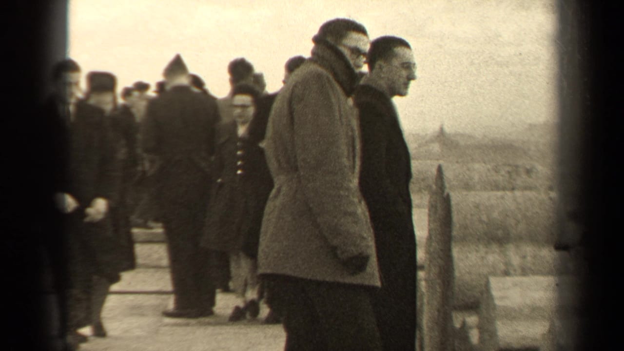 People in heavy coats stand and walk on a paved overlook, with two men prominent at right.