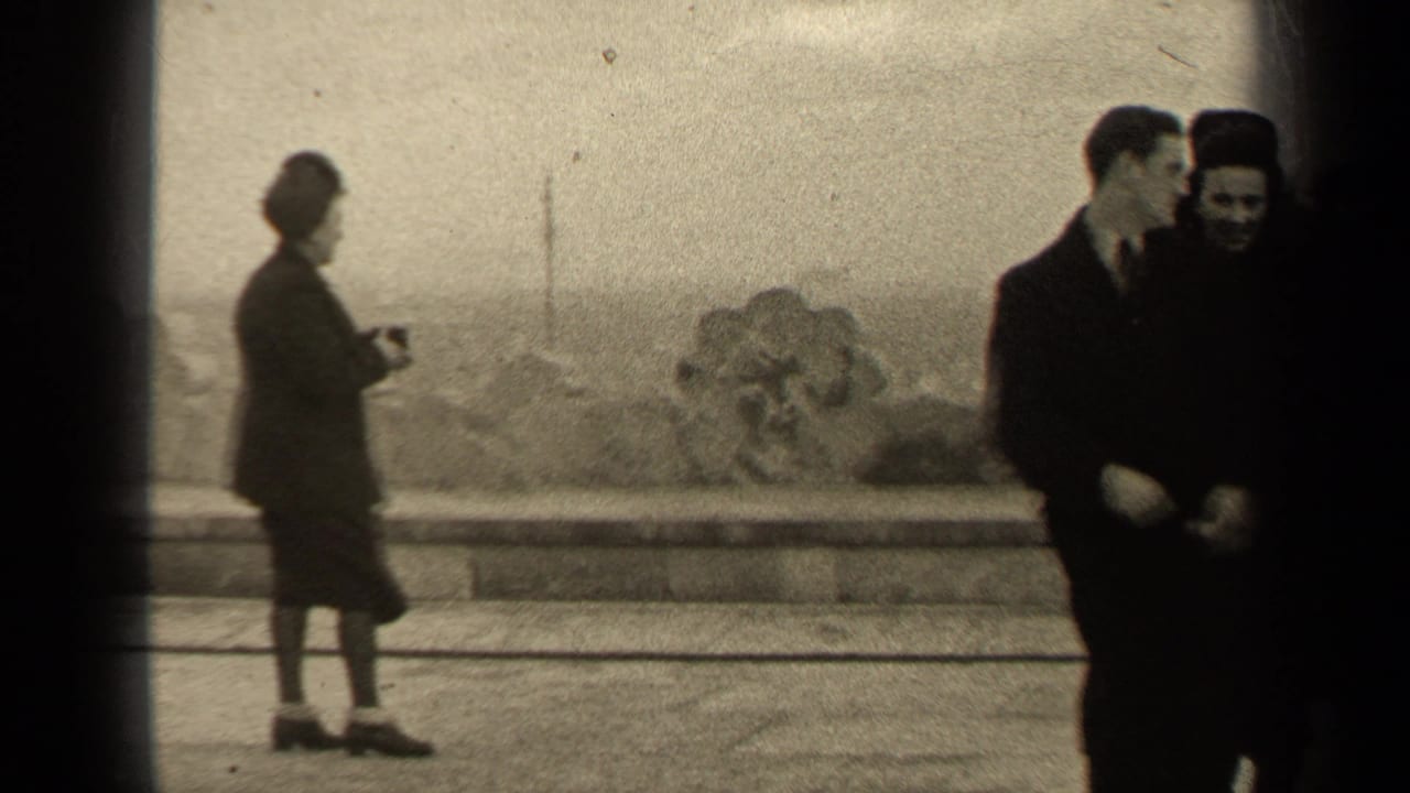 Adults in dark clothing stand near a stone balustrade while a woman holds a camera.