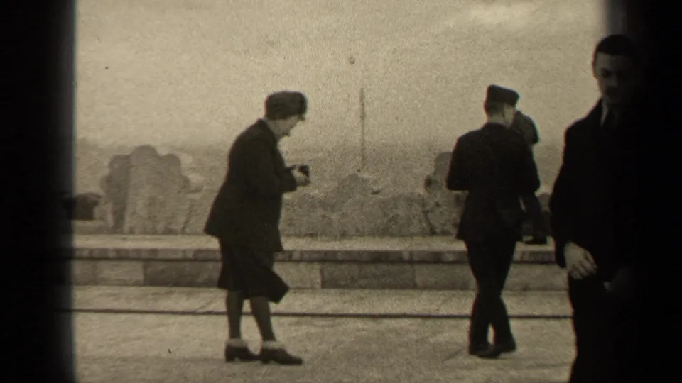 1947 Paris frame with a woman holding a camera, a uniformed man, and formally dressed adults beside a stone barrier.
