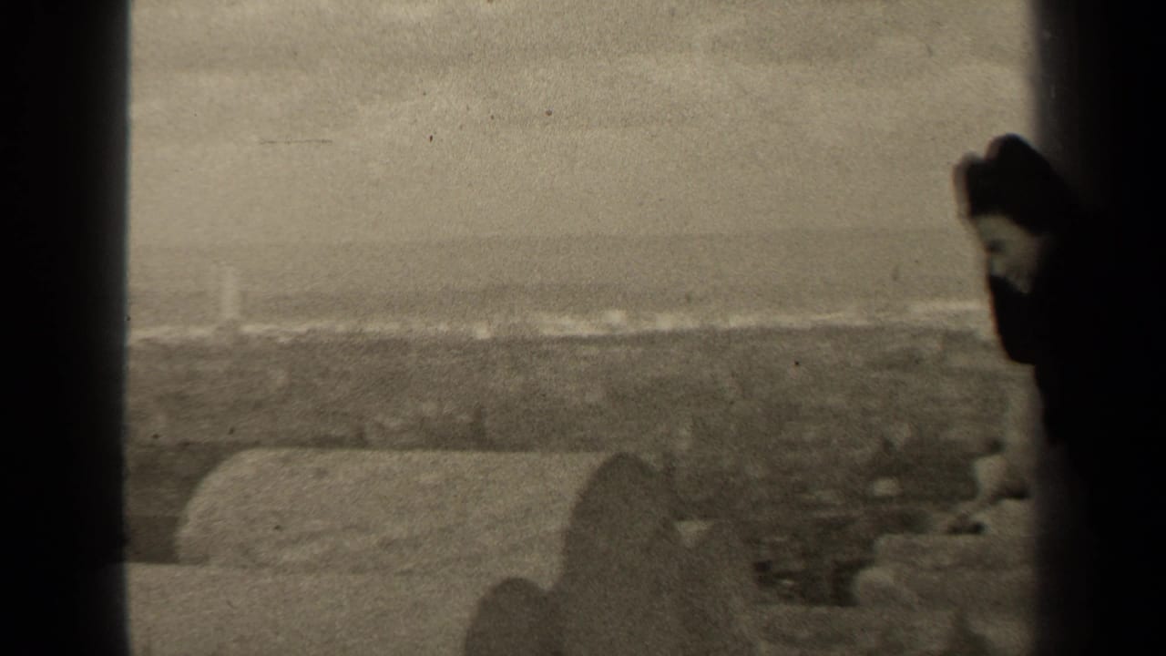A dark-clad woman appears at the right edge with rooftops and horizon behind her.